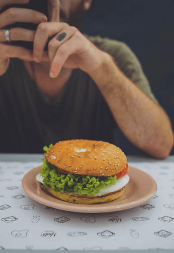 Photo by Toa Heftiba on Unsplash The image shows a bagel with salad, tomatoes and mozzarella cheese on a plate on a table. In the background a person is sitting at the table, looking at a mobile phone.